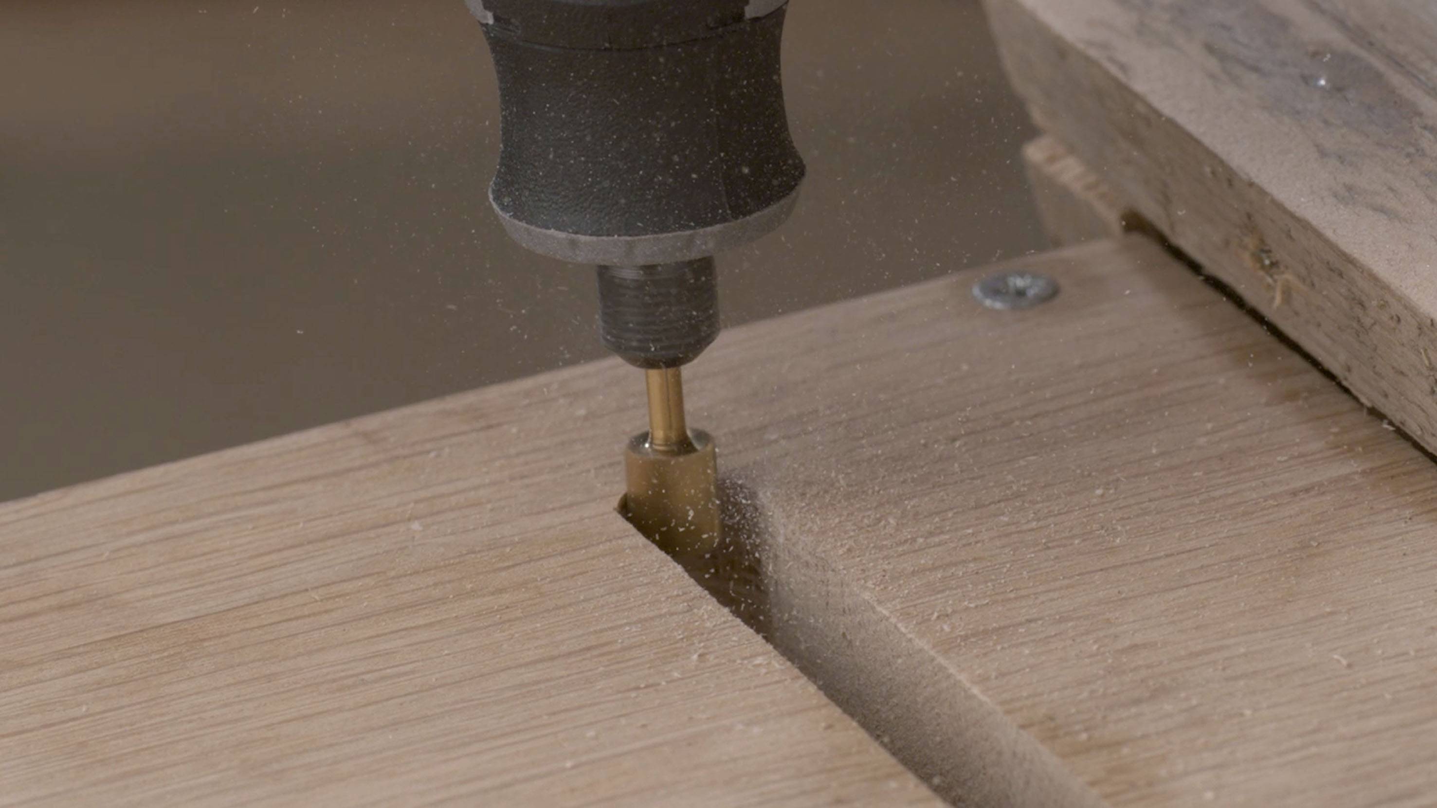 Detailní záběr frézky opracovávající dřevěnou desku, při které vzniká pilina.