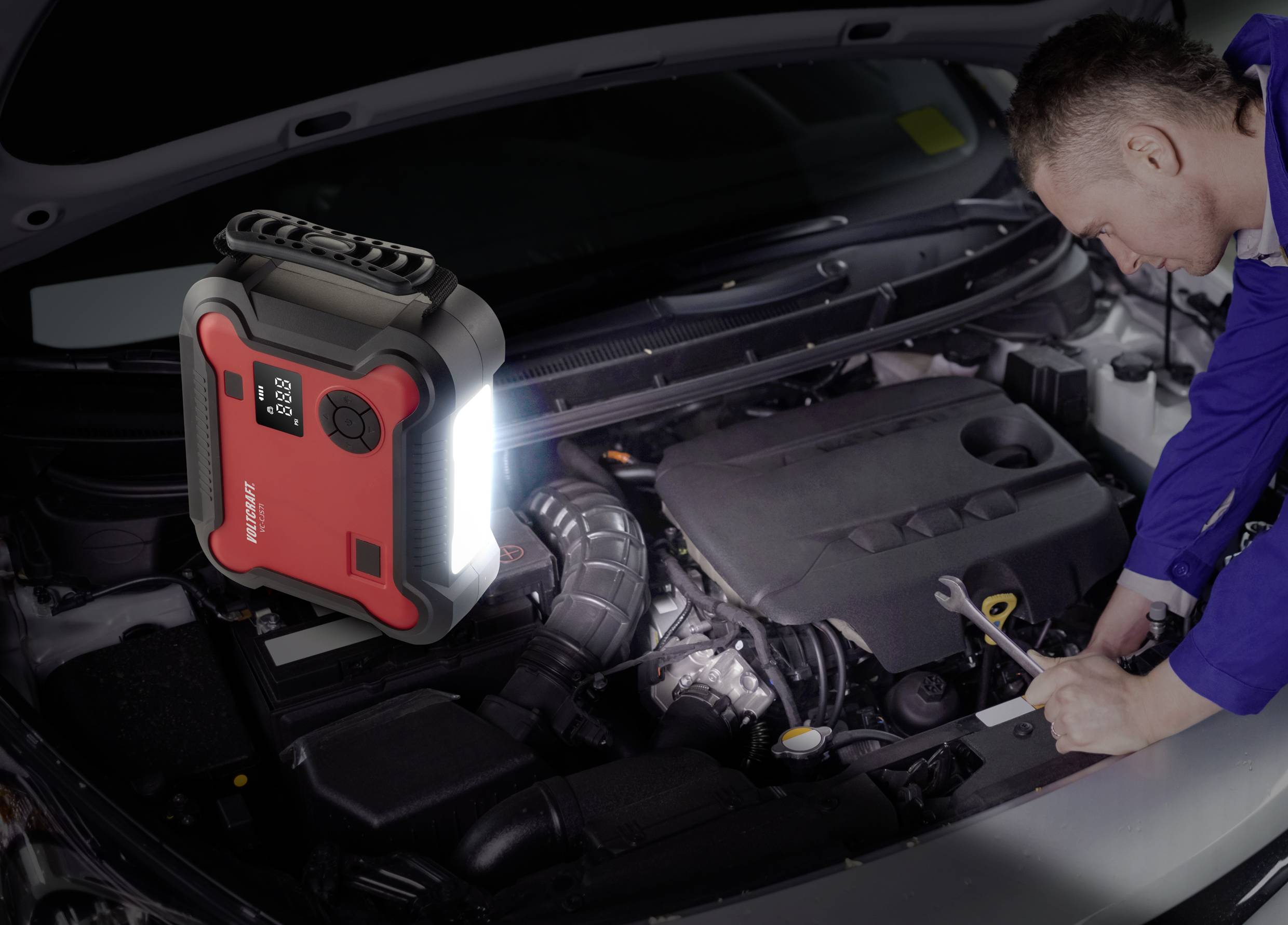 Muž pracuje s klíčem na motoru automobilu; v popředí červený startovací pomocný přístroj s vestavěným světlem.