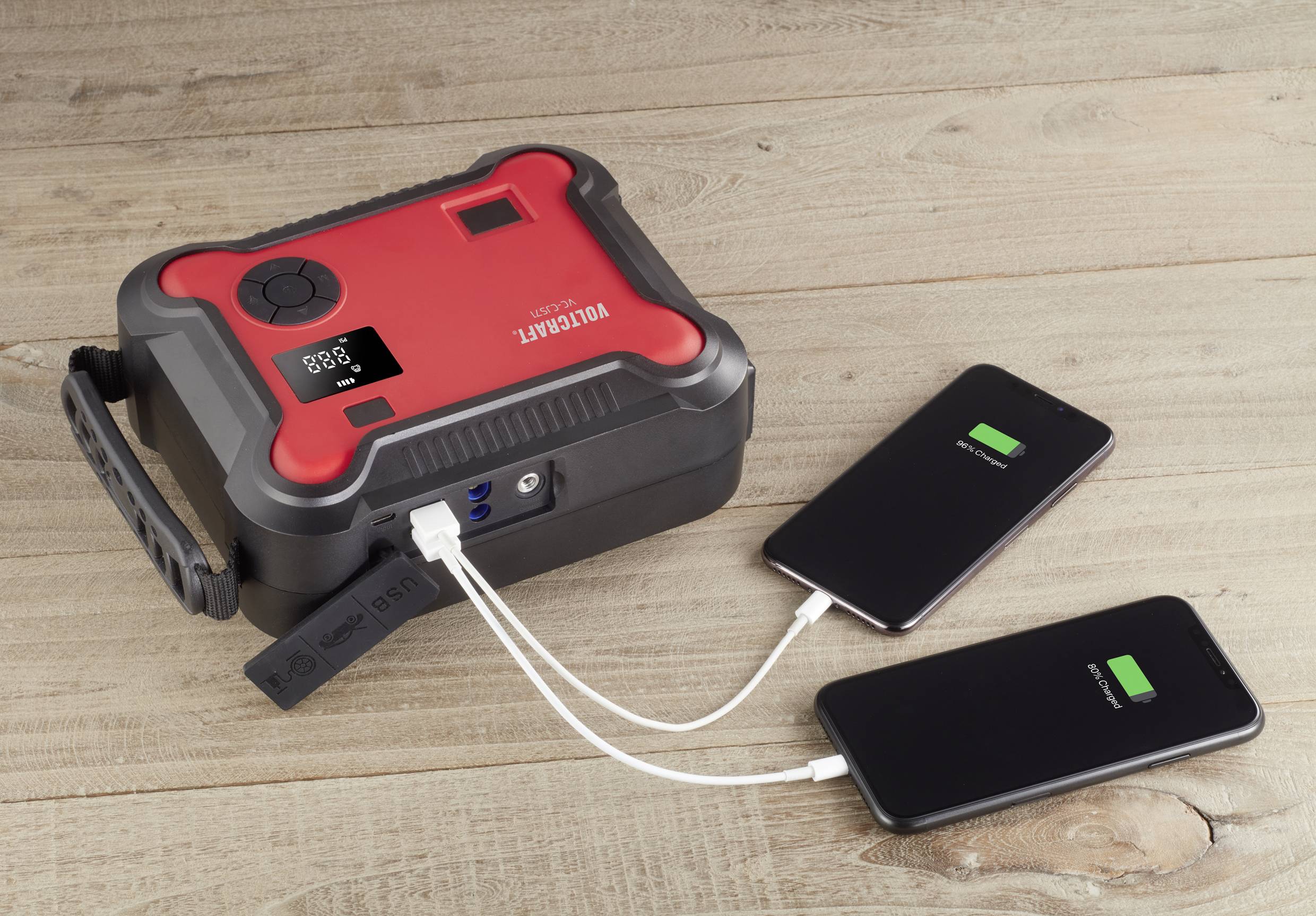 Přenosná powerbanka na dřevěném stole nabíjí dva smartphony pomocí USB kabelu, přičemž oba zobrazují zelený stav nabití baterie.