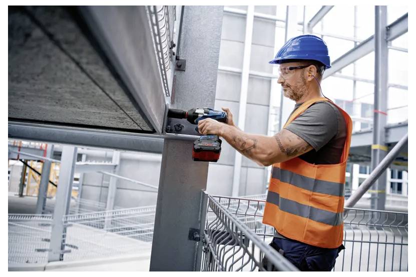 Stavební dělník v modré ochranné přilbě a oranžové vestě používá elektrickou vrtačku na ocelové nosníky uvnitř budovy, která je ve výstavbě.