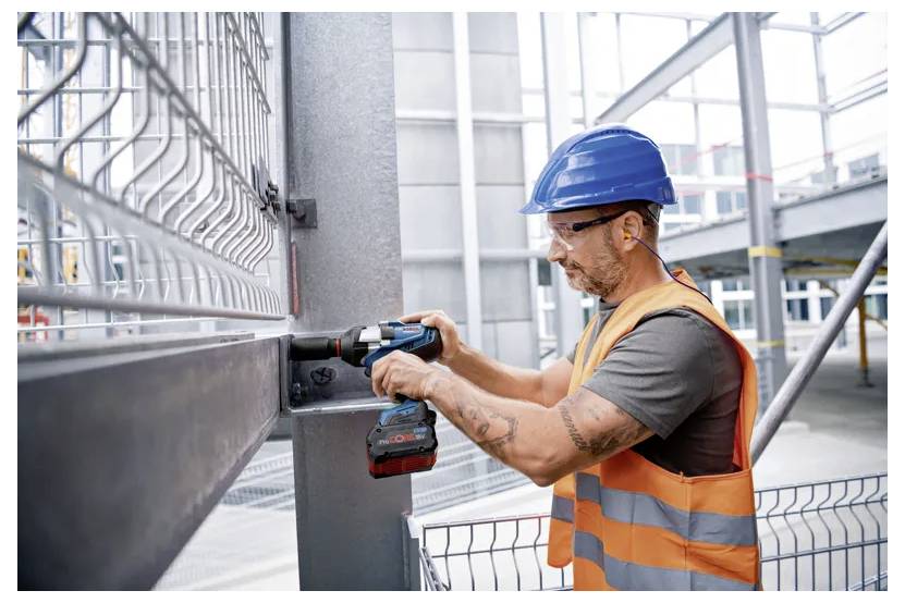 Dělník ve stavebním průmyslu v modré přilbě a oranžové vestě používá elektrickou vrtačku k upevnění kovového rámu na staveništi.