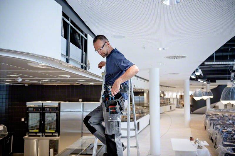 Technik opravuje něco na žebříku v moderním, otevřeném prostoru. V pozadí jsou vidět kuchyňské spotřebiče.