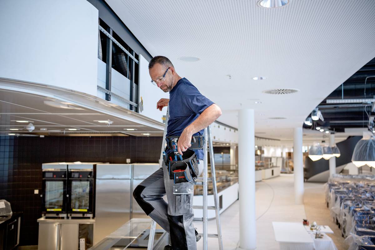 Řemeslník na žebříku opravuje stropní svítidlo v moderní restauraci s několika sedacími zónami.