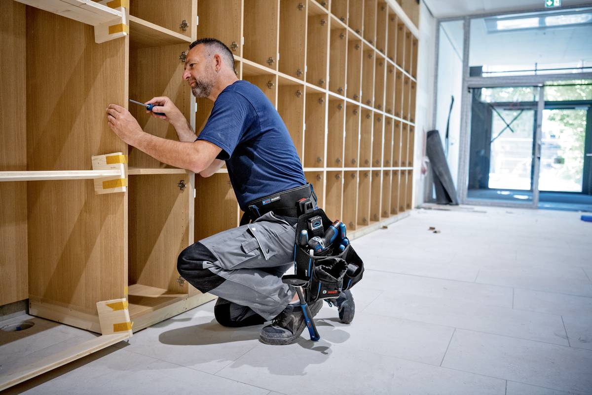 Řemeslník v modrém oblečení montuje police v místnosti. Klečí na podlaze a drží v ruce nářadí.