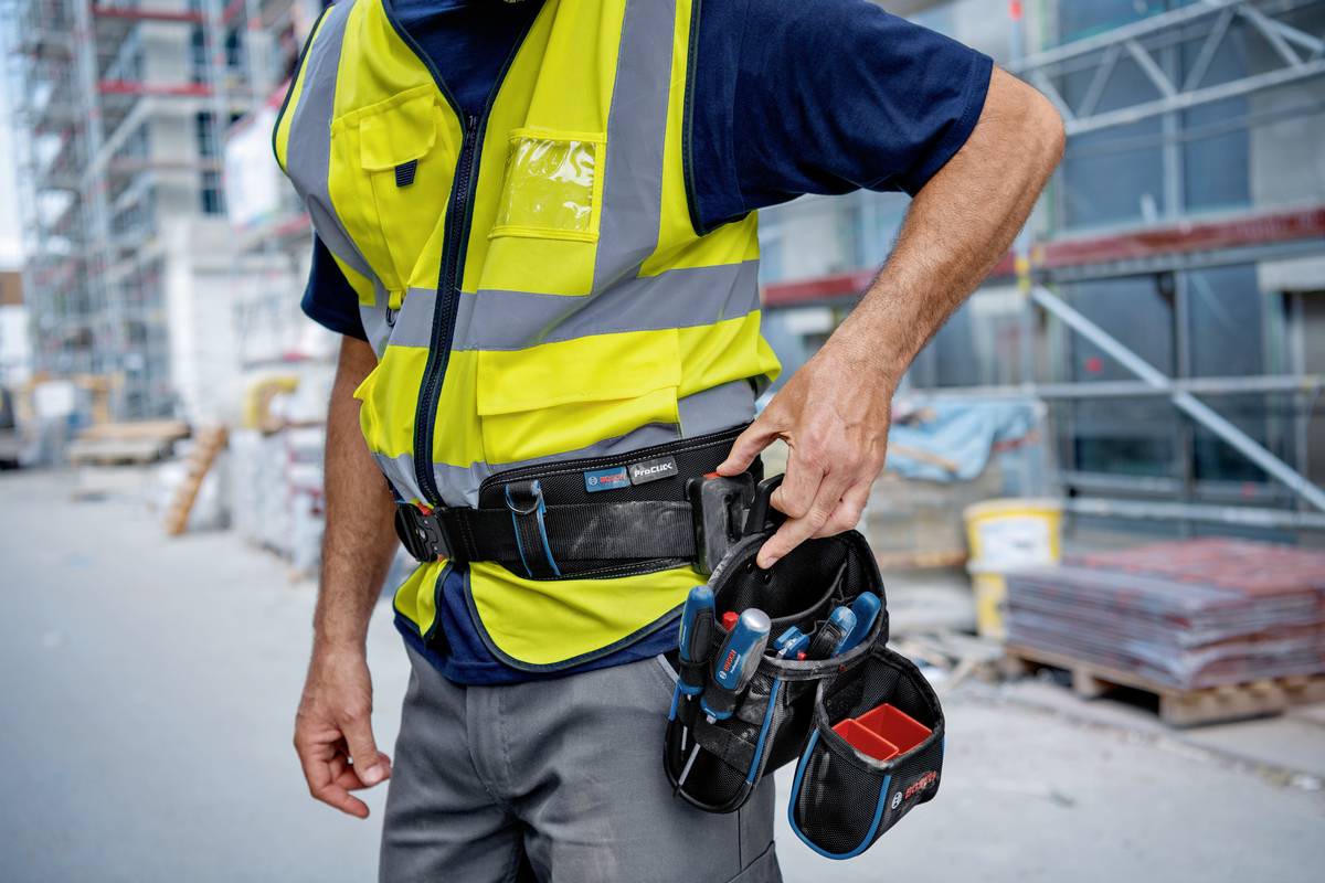 Dělník na staveništi je oblečen v reflexní bezpečnostní vestě a má opasek na nářadí. V pozadí jsou lešení a stavební materiály.