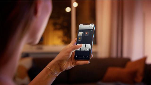 Osoba drží smartphone a pomocí aplikace ovládá osvětlení v útulném, teplým světlem osvětleném obývacím pokoji.