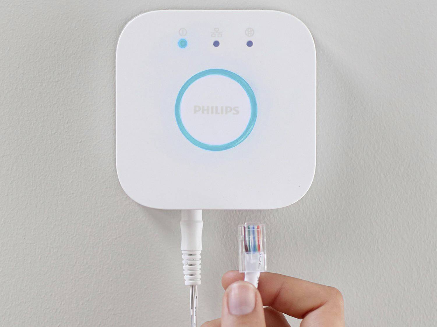 Osoba drží síťový konektor blízko chytré domácí brány Philips se svítícím modrým prstencem a logem 'Philips'.