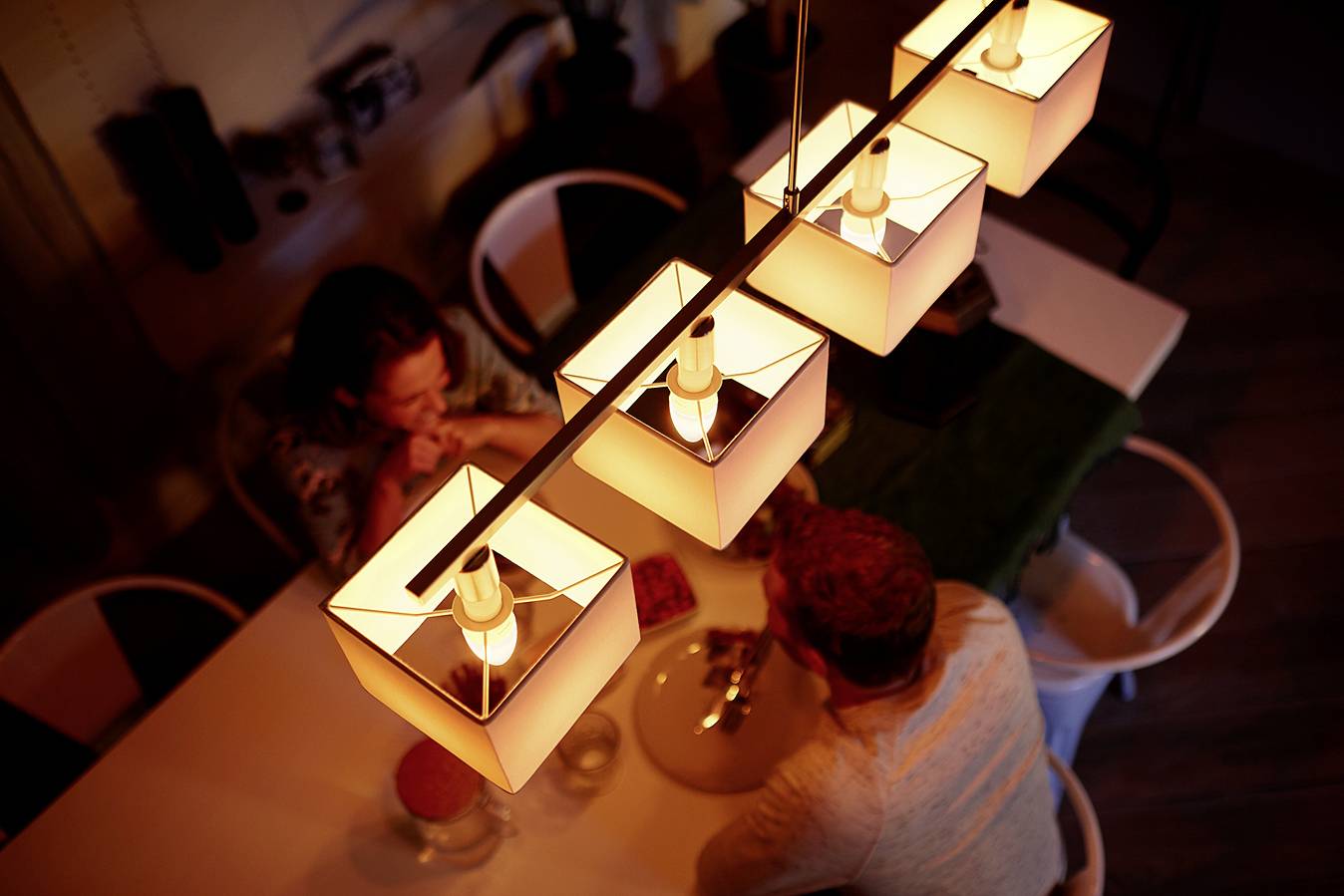 Pár sedí u stolu pod řadou pěti zářících lamp. Atmosféra je teplá a útulná.