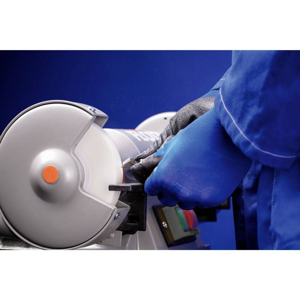 Osoba v modré pracovní kombinéze obsluhuje brusku a přitiskává nástroj k rotujícímu kotouči.