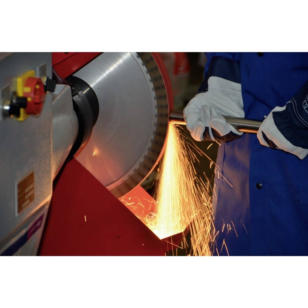 Osoba v modré pracovní kombinéze brousí kov na rotující stroji, přičemž z místa broušení odlétávají jiskry.