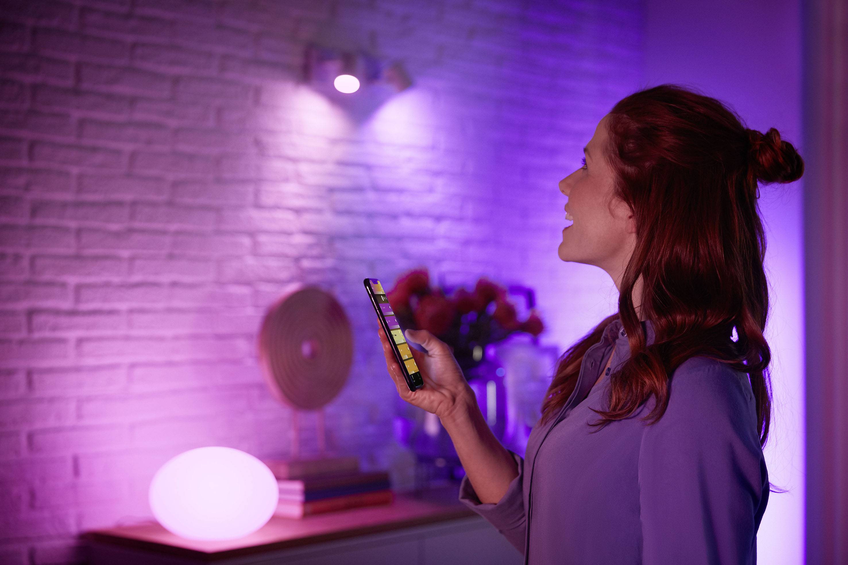 Žena ovládá barevné osvětlení smartphonem v atmosféricky osvětlené místnosti.