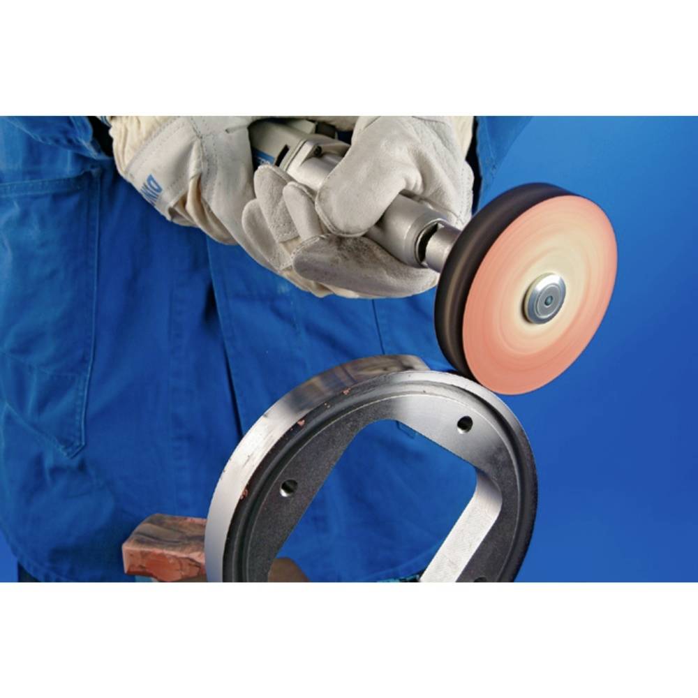 Dělník v modré kombinéze a rukavicích brousí kovový díl pomocí rotující brusné kotouče.