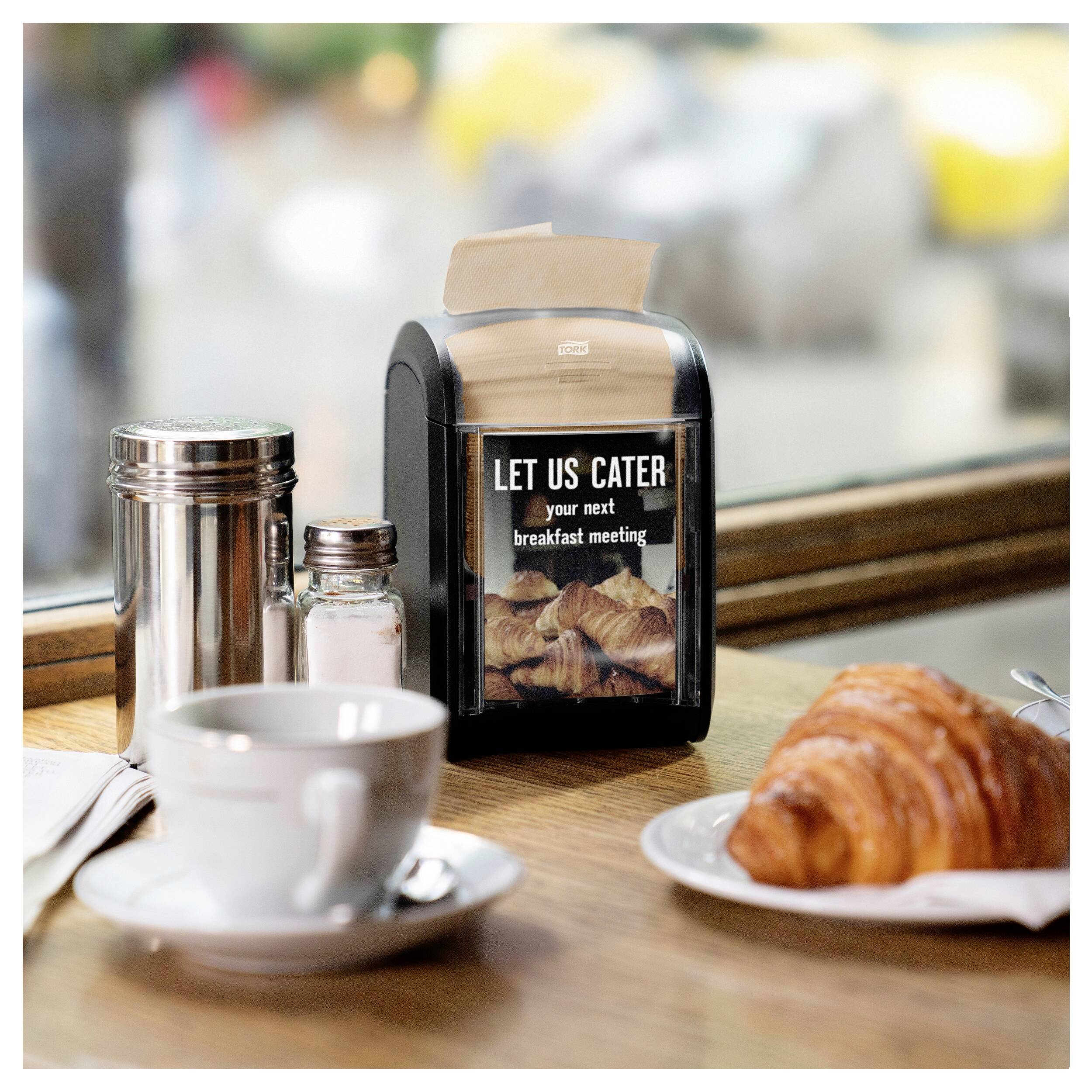 Na stole stojí šálek, croissant a papírový držák s nápisem 'Zajistíme catering pro vaše příští snídaňové setkání'.