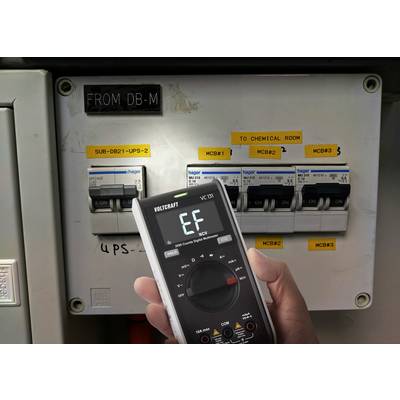Digitální multimetr zobrazuje 'EF', zatímco je namířen na rozvodnou skříň s několika pojistkami. Štítky ukazují různé elektrické okruhy.