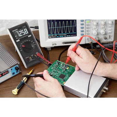 Osoba testuje zelenou elektronickou desku plošných spojů multimetrem, který leží na stole. V pozadí je zobrazení na osciloskopické obrazovce.