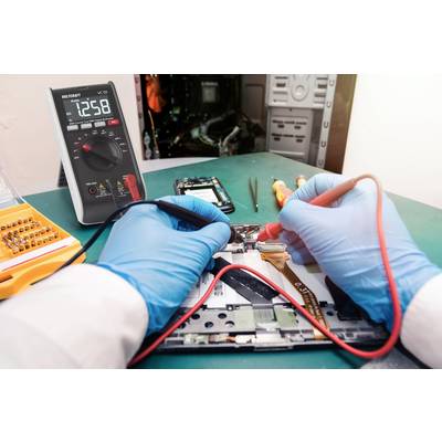 Osoba v modrých rukavicích opravuje elektronické zařízení na stole. V pozadí je multimetr s displejem zobrazujícím '12.58'.