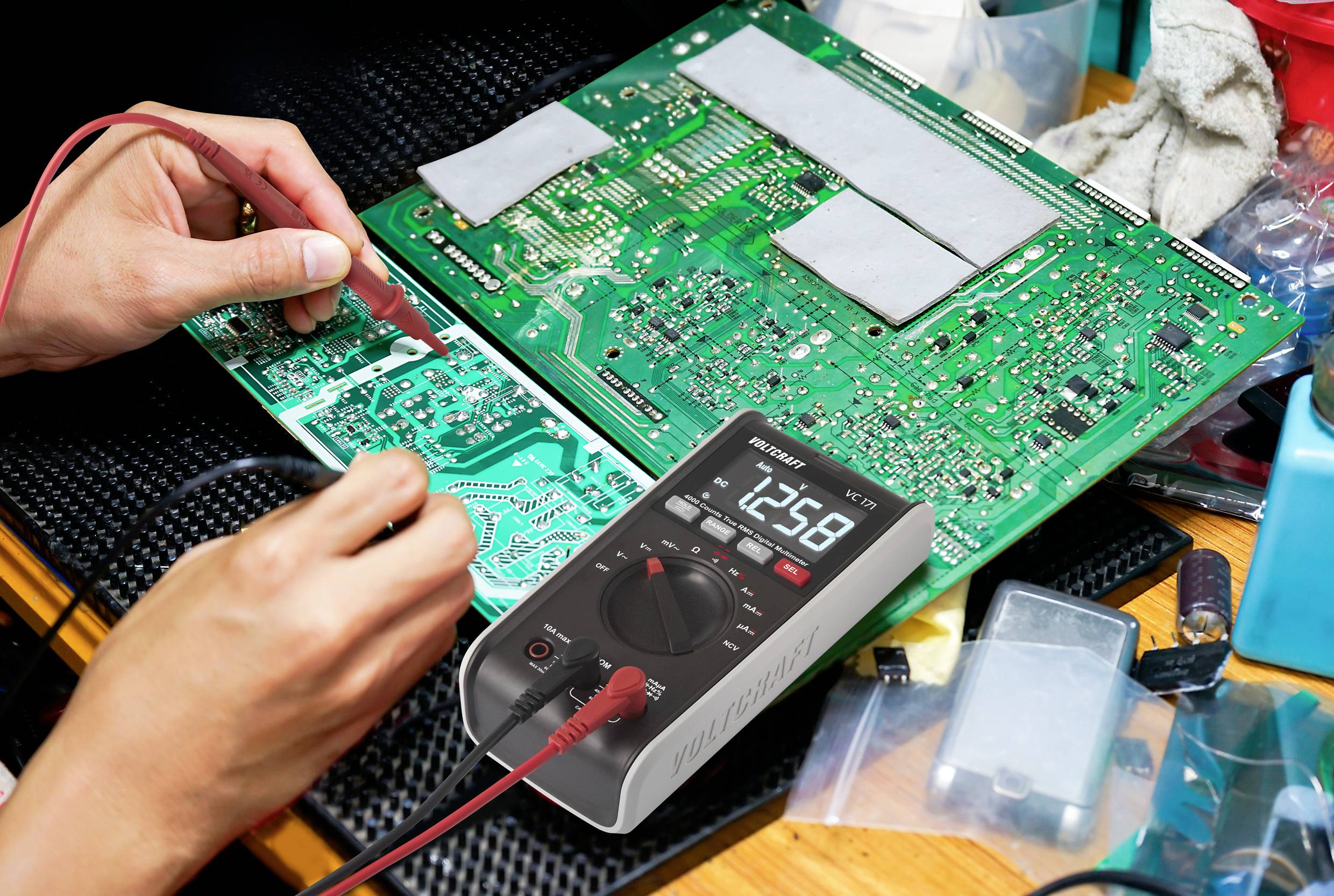 Osoba testuje zelenou desku plošných spojů multimetrem. Displej ukazuje '12.58'. Obklopena elektronikou a nástroji.