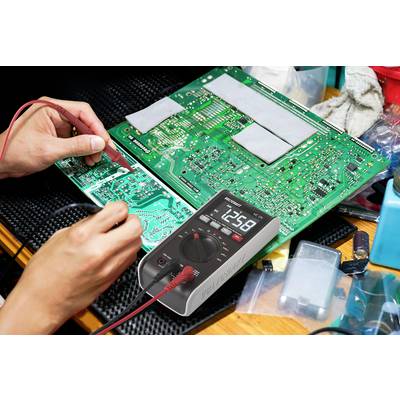 Osoba testuje zelenou desku plošných spojů multimetrem. Displej ukazuje '12.58'. Obklopena elektronikou a nástroji.
