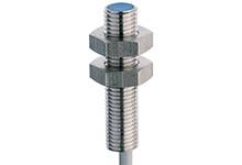 Kovový senzor s válcovým pouzdrem, závitem a kabelovým připojením, používaný k detekci kovových objektů v průmyslových aplikacích.