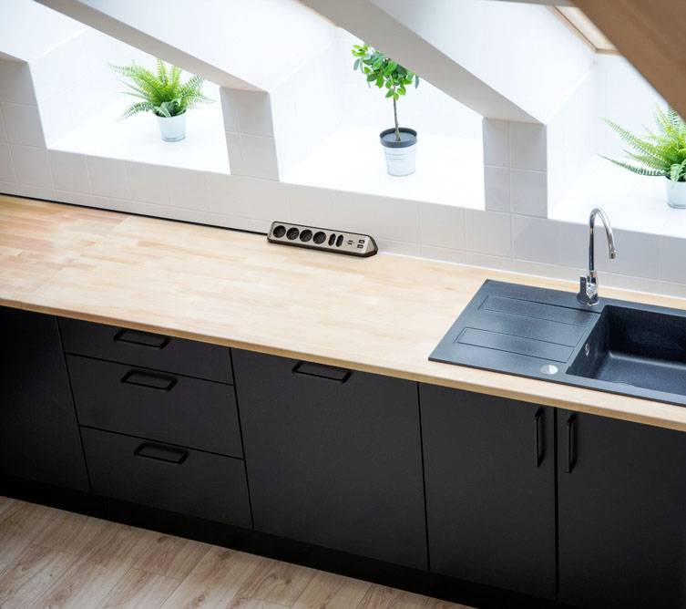 Dřevěná kuchyňská pracovní deska s černým dřezem, šestinásobná zásuvková lišta a tři rostliny před velkým oknem pod šikmou střechou.