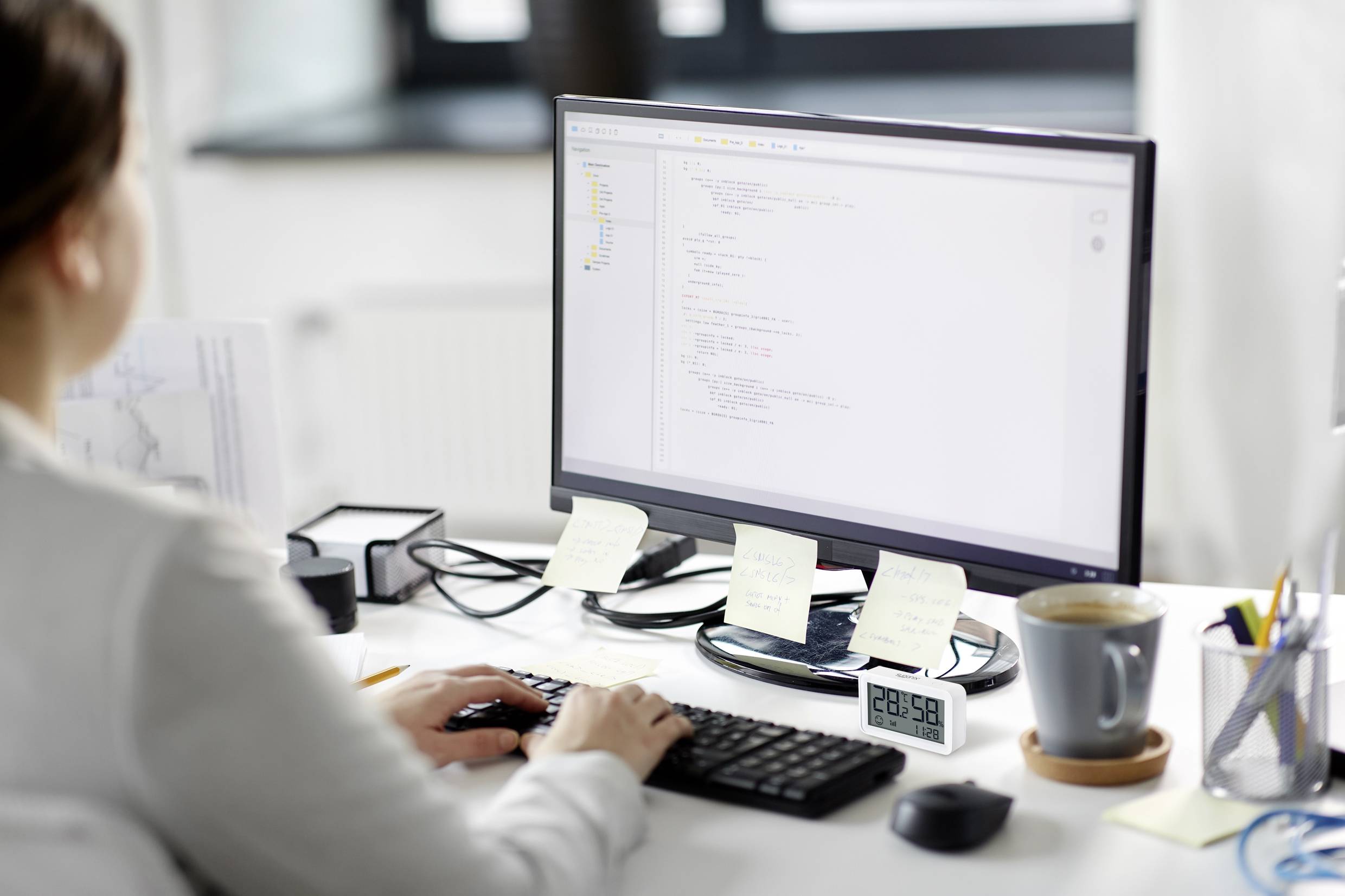 Osoba pracuje u psacího stolu před monitorem zobrazujícím programátorský kód. Na stole stojí hrnek a leží poznámky.
