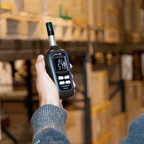 Osoba drží měřicí přístroj, který ukazuje teplotu 5,7 stupňů Celsia. V pozadí jsou viditelné regály s krabicemi.