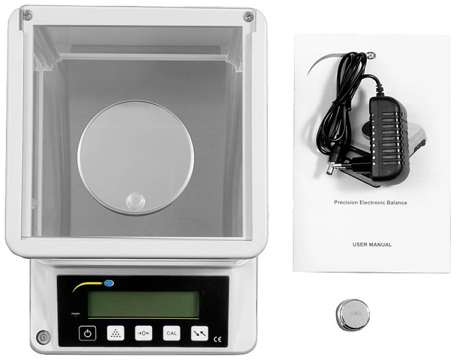 Elektronická přesná váha s průhledným krytem, návod k obsluze, napájecí adaptér a kalibrační závaží vedle ní.