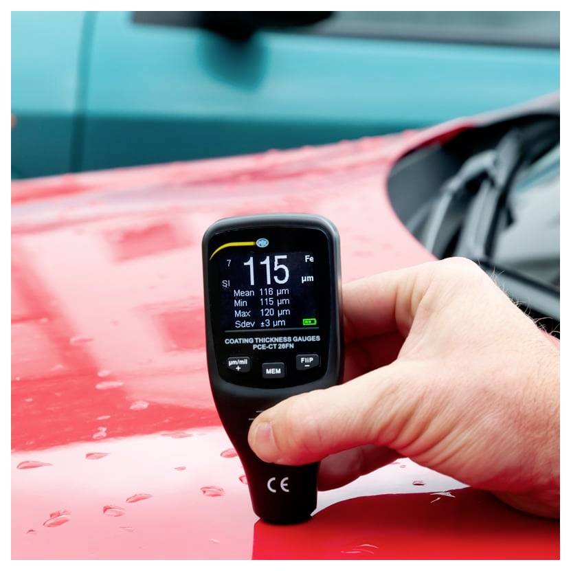 Přenosný měřič tloušťky nátěru měří barvu na červené kapotě automobilu a na jeho displeji zobrazuje 115 µm.