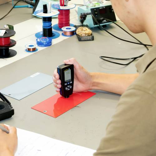 Člověk měří tloušťku červeného materiálu digitálním měřicím přístrojem na pracovním stole s nástroji a kabelovými svazky.