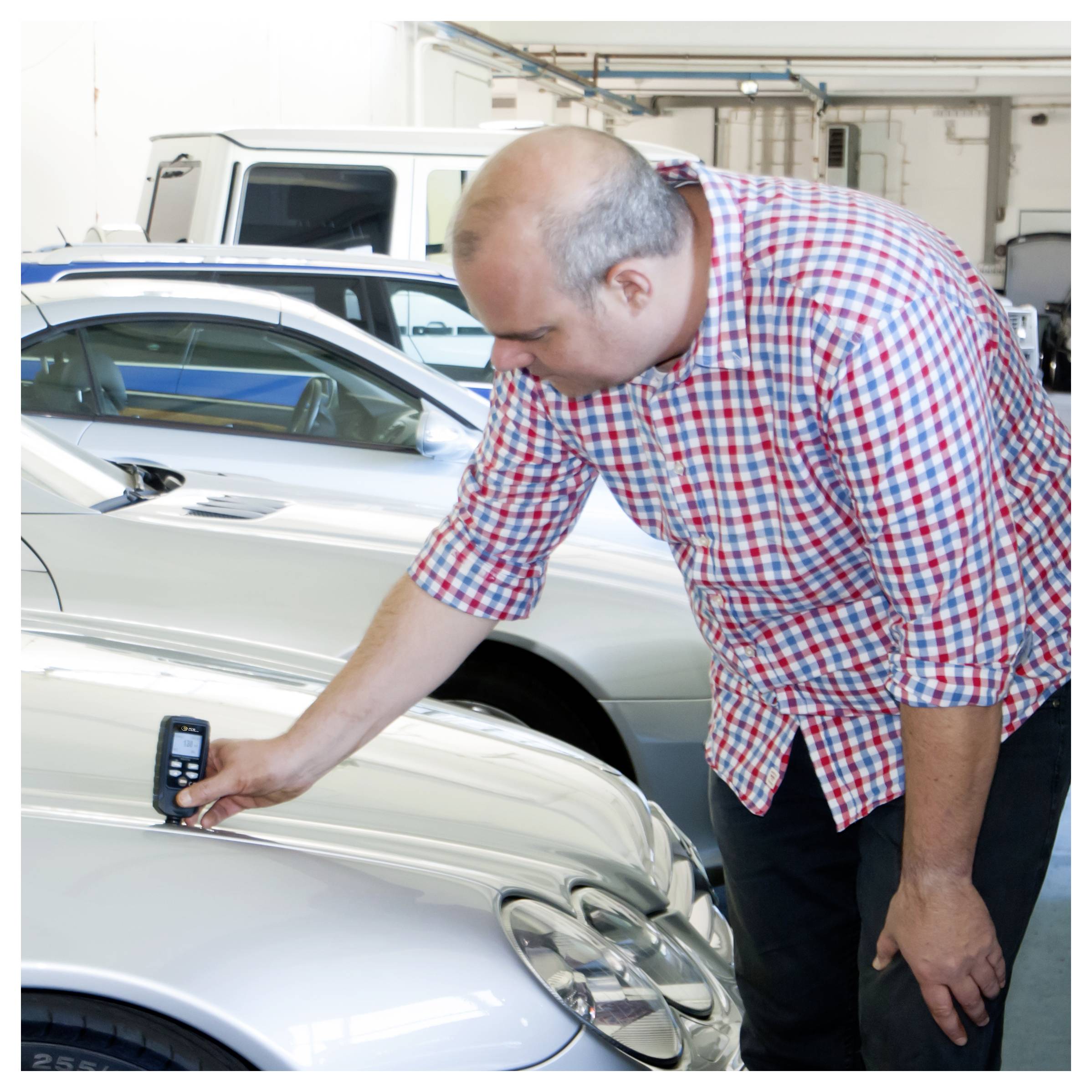 Muž inspekuje přední část stříbrného automobilu v garáži a zdá se, že používá ruční zařízení na kapotě.