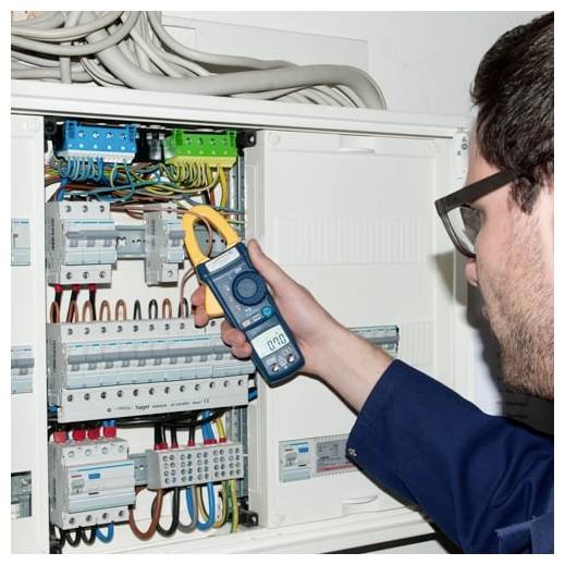Technik testující elektrický panel kleštovým ampérmetrem, měřící vedení uvnitř otevřené skříně jističů.