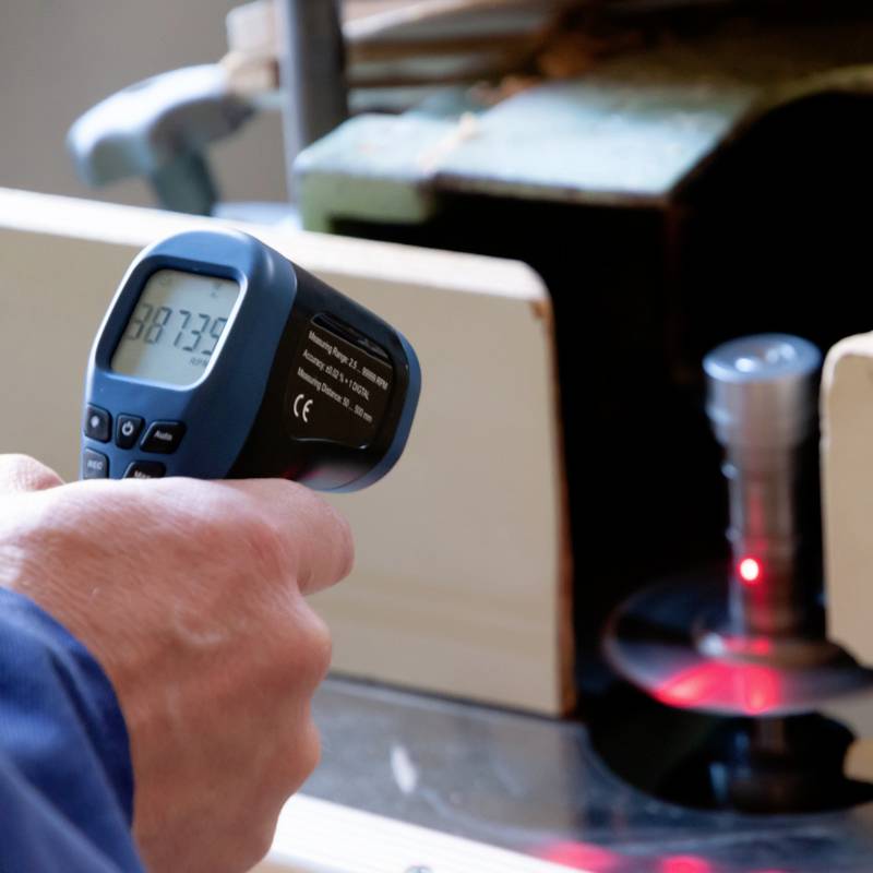 Osoba drží infračervený teploměr a odečítá teplotu 87,3 °C, zjevně za účelem měření stroje.