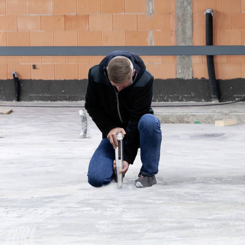 Osoba klečí na betonovém podlaze a používá ruční nástroj. V pozadí je vidět oranžová stěna a potrubí.