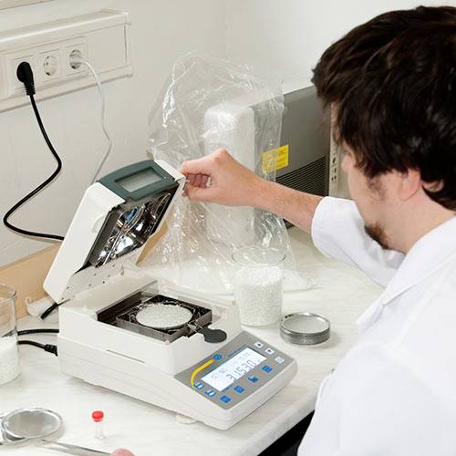 Vědec v laboratoři otevírá analytický přístroj s vzorky, obklopen elektrickými zásuvkami a laboratorním vybavením.