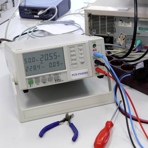 Digitální ampérmetr ukazuje '20,55' při '220 V AC'. Vedle něj leží šroubovák a kleště. Pozadí tvoří laboratorní prostředí.