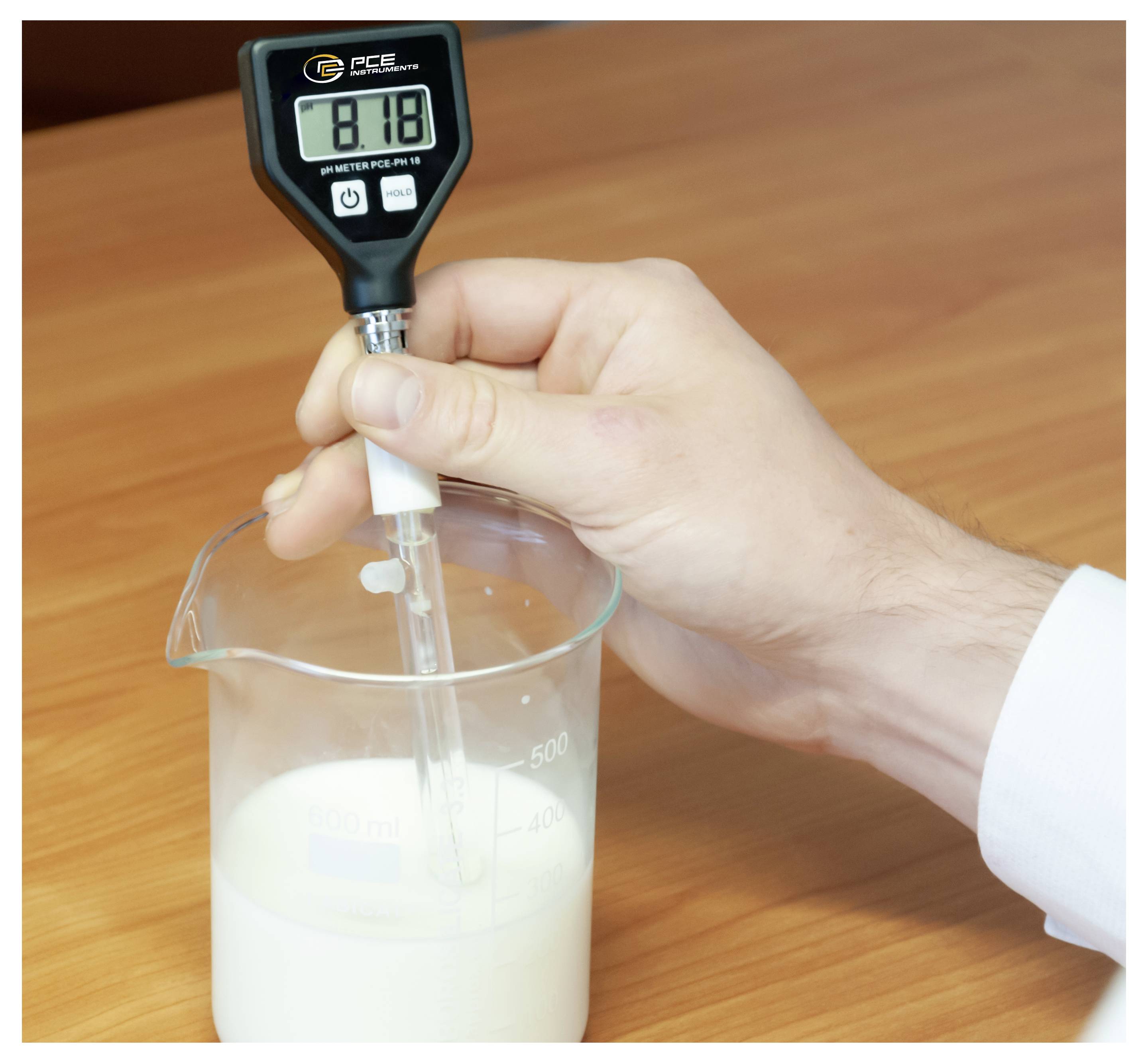 Ruka držící digitální pH metr v kádinovi s mlékem na dřevěném stole, měření acidity.