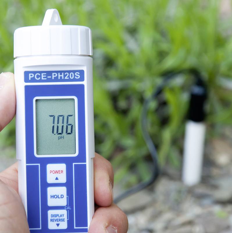 Jedna ruka drží pH metr s displejem ukazujícím '7,06 pH'. V pozadí je viditelná tráva.