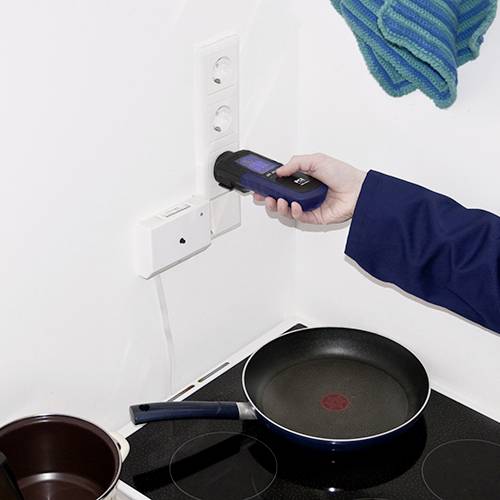 Osoba v modré bundě drží měřič spotřeby energie zasunutý do elektrické zásuvky. Vedle toho stojí hrnec na sporáku.