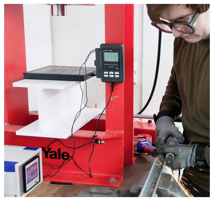 Technik používá elektrické nářadí k testování montáže zámku Yale namontované v červeném laboratorním rámu s připojenými digitálními displejemi měření.