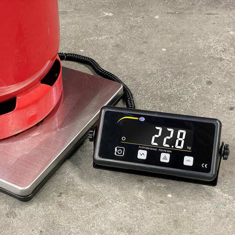 Červená plynová láhev stojí na digitální váze, která ukazuje 22,8 kg. Váha a láhev se nacházejí na šedé podlaze.