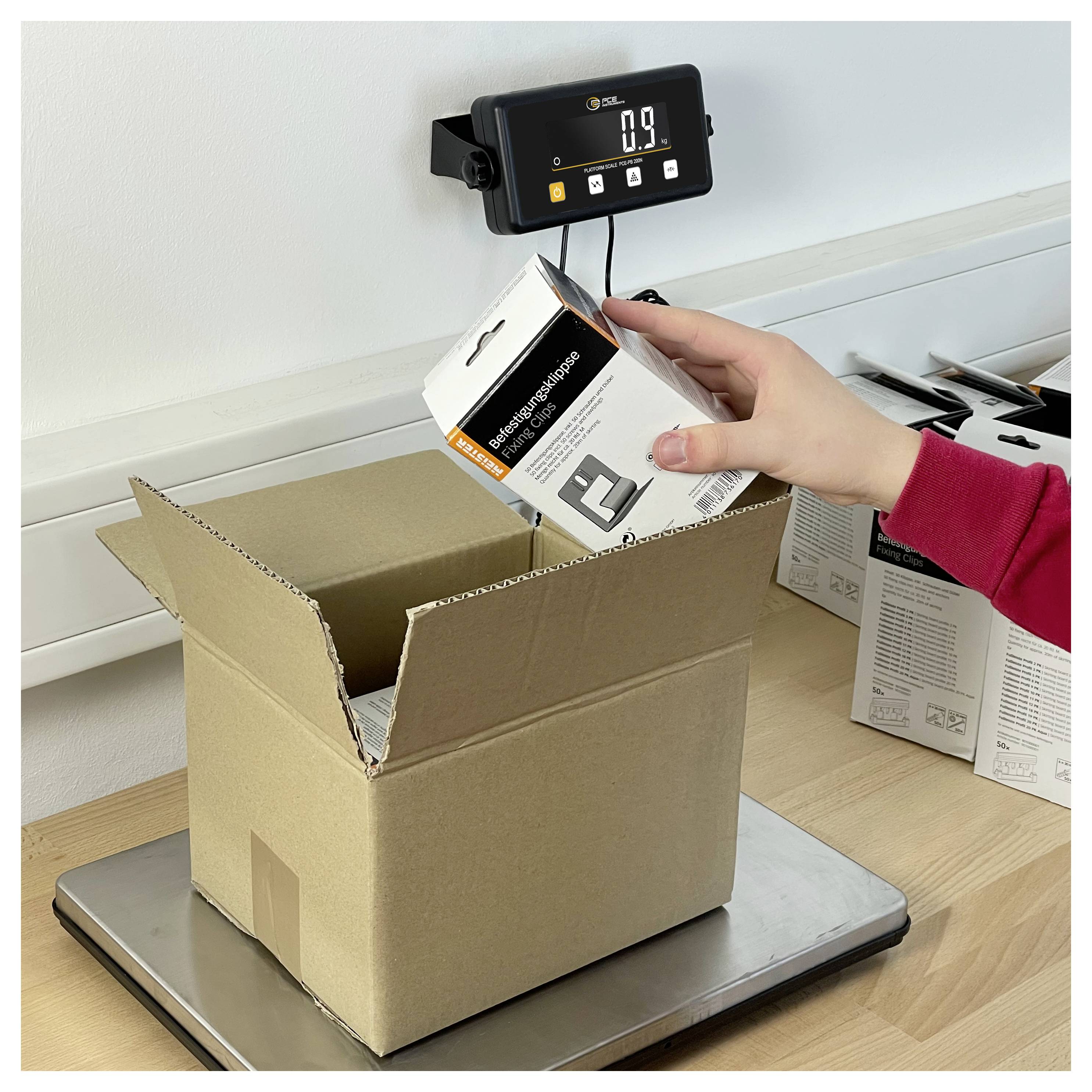 Osoba umisťuje maloobchodní krabici s produktem do cardboardové přepravní krabice na digitální váze, která ukazuje 0,9 kg.