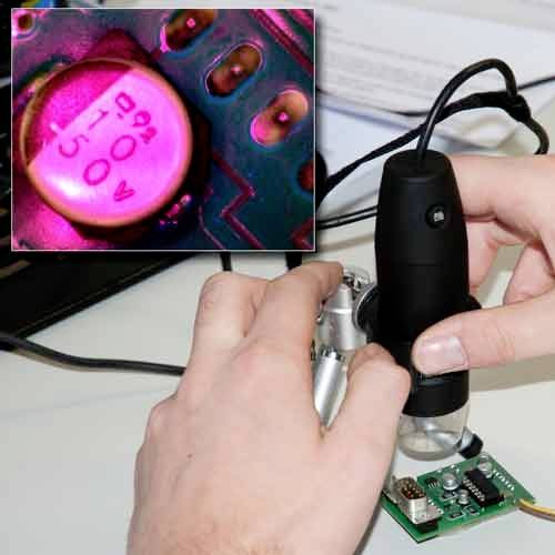 Osoba zkoumá elektronické součástky pomocí mikroskopu. Detailní záběr ukazuje čísla na kondenzátoru.