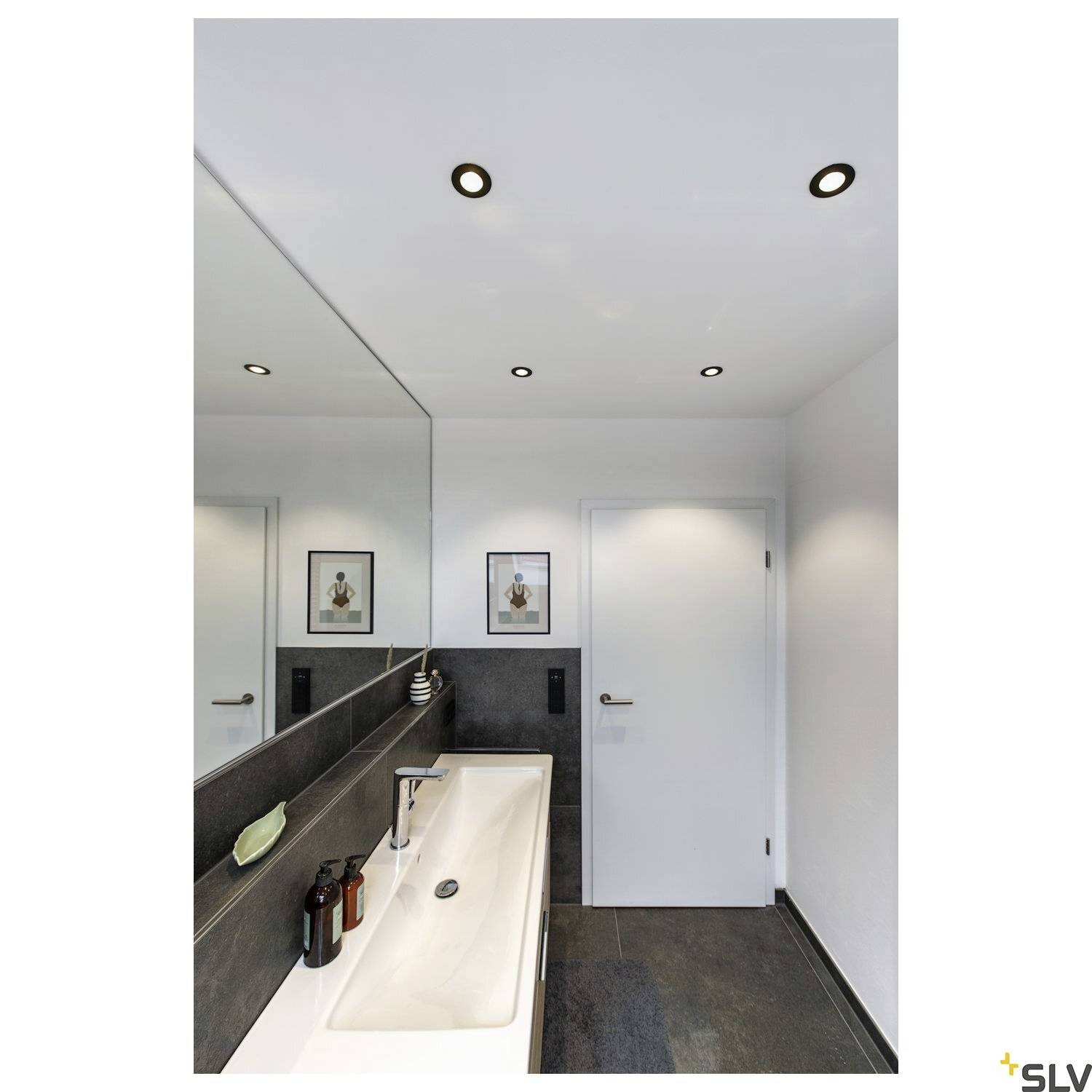 Moderní koupelna s obdélníkovým umyvadlem, zrcadlem nad ním, dvěma černými obrazovými rámy na bílé stěně a tmavou dlažbou.