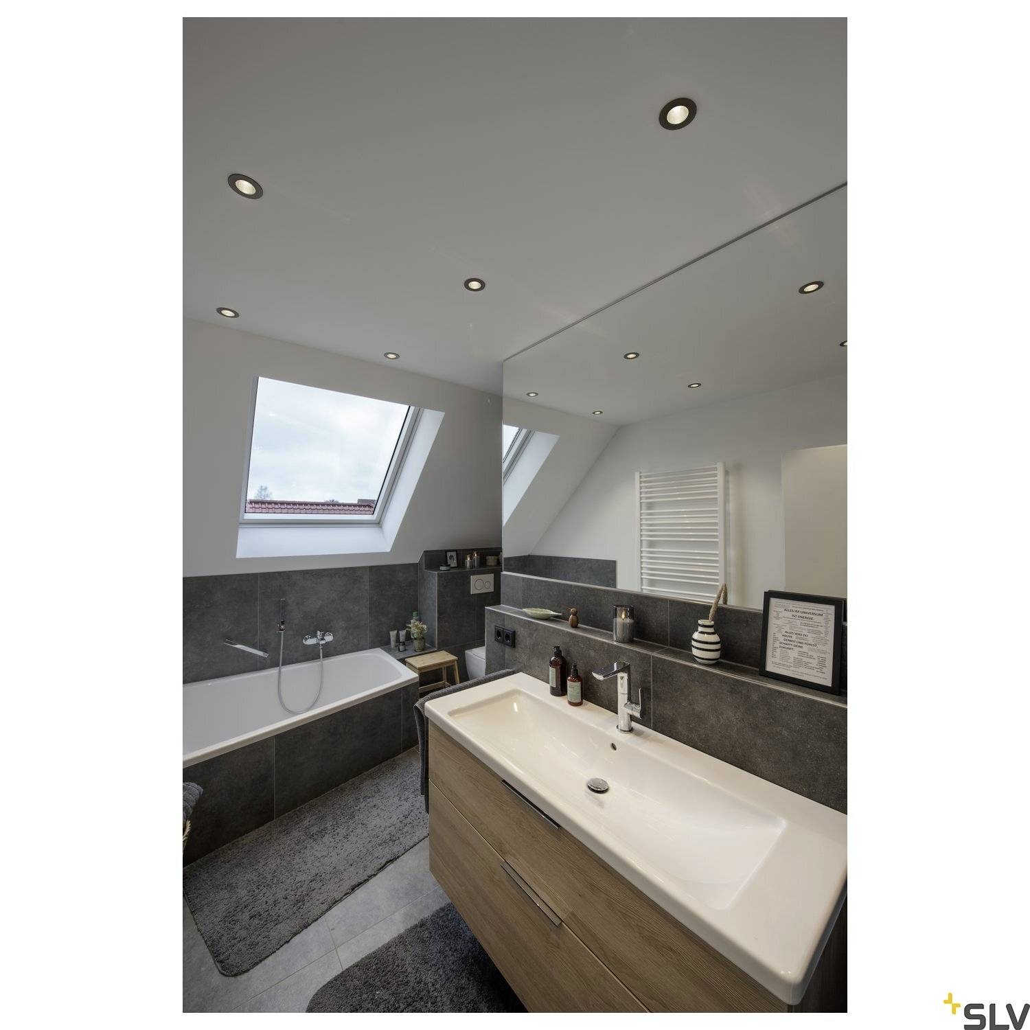 Moderní koupelna se střešním oknem. Vlevo vana, vpravo umyvadlo se zrcadlem. Světlé nástěnné osvětlení. Elegantní, minimalistický design.