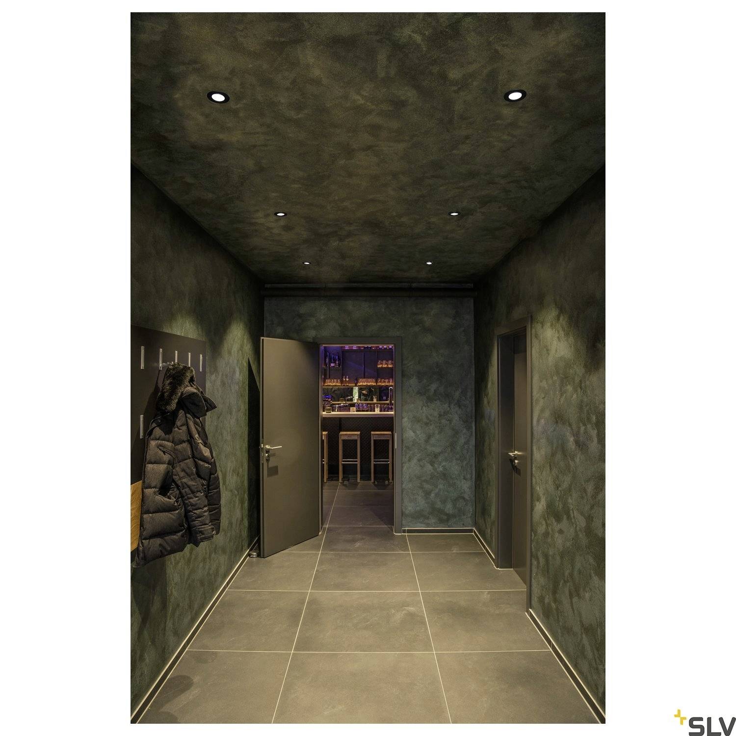 Chodba s tlumeným osvětlením, vlevo visí bunda. Otevřené dveře odhalují pohled do baru s osvětleným barem.