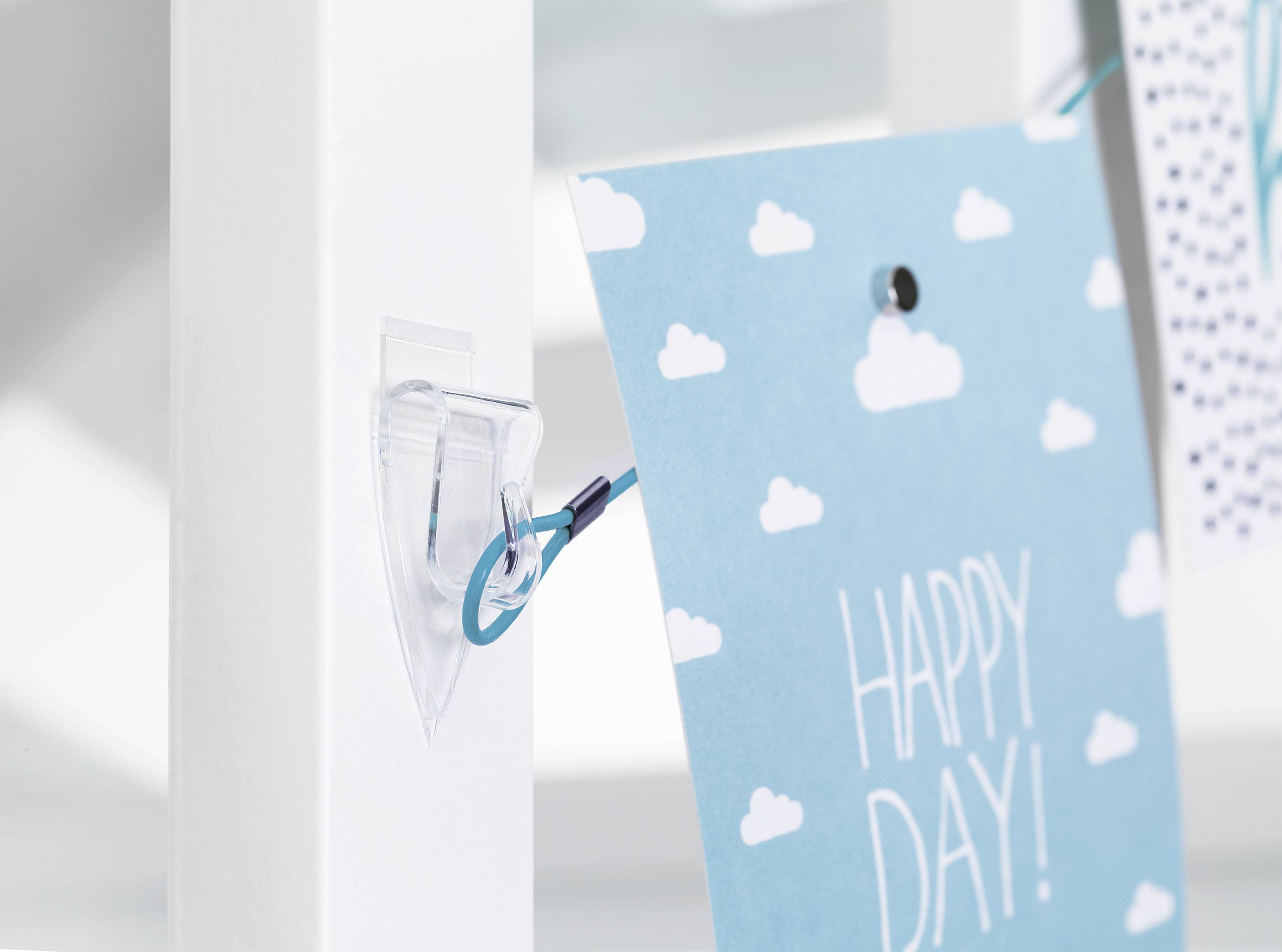 Pohlednice s bílými mraky a textem 'HAPPY DAY!' je připevněna modrou stuhou na bílé vertikální liště.