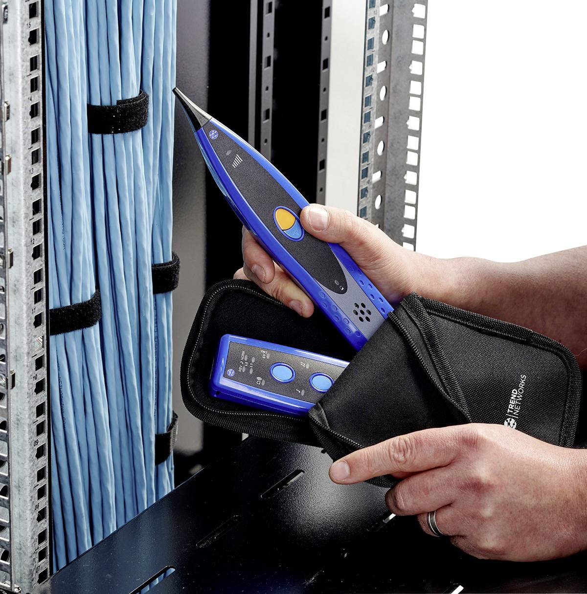 Osoba drží modré testovací zařízení pro kabely vedle otevřeného serverového racku s modrými kabely, připravená na síťovou kontrolu.