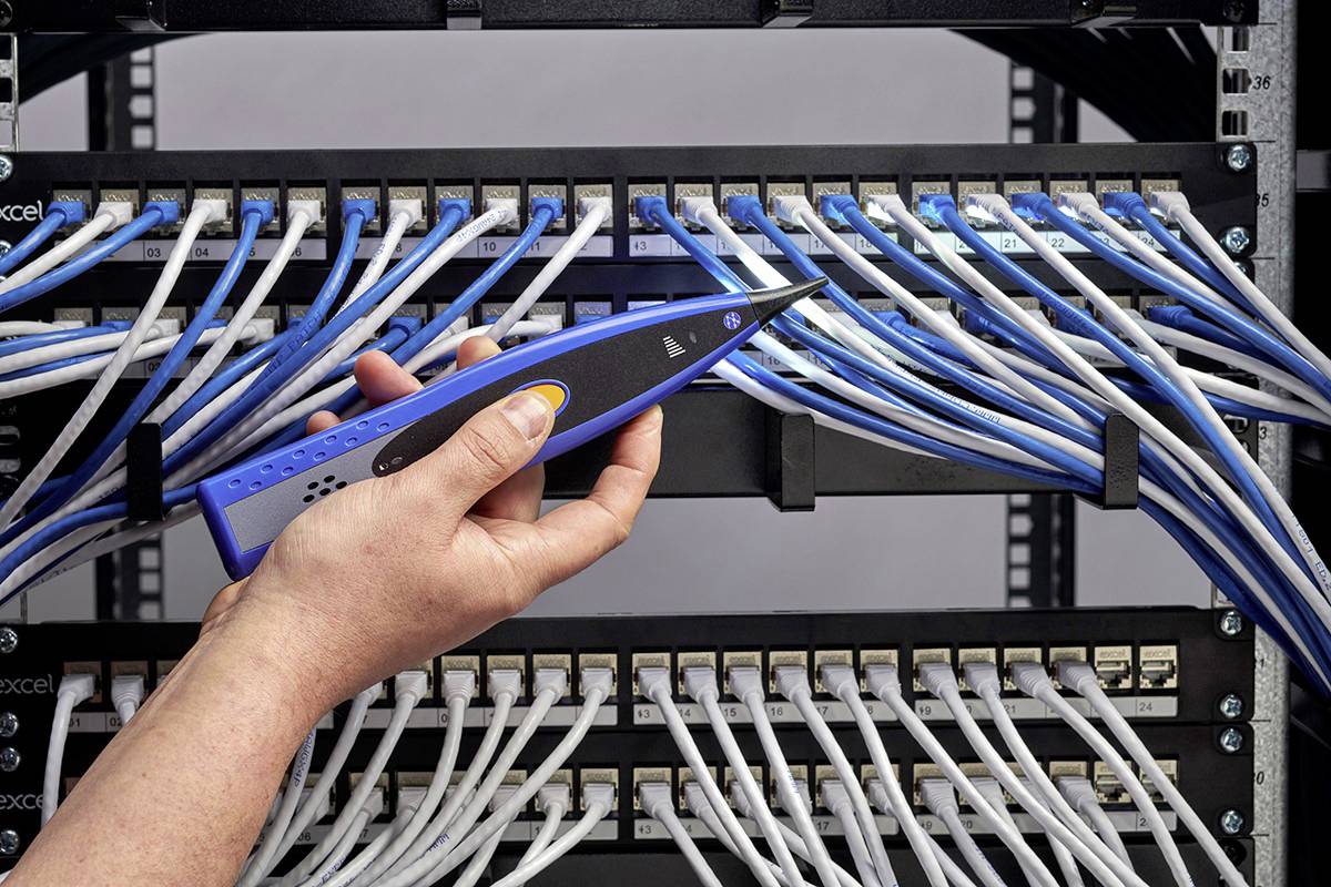 Jedna ruka drží testovací zařízení připojené k elektrickým kabelům v serverovně. Různé kabely propojují síťové komponenty.