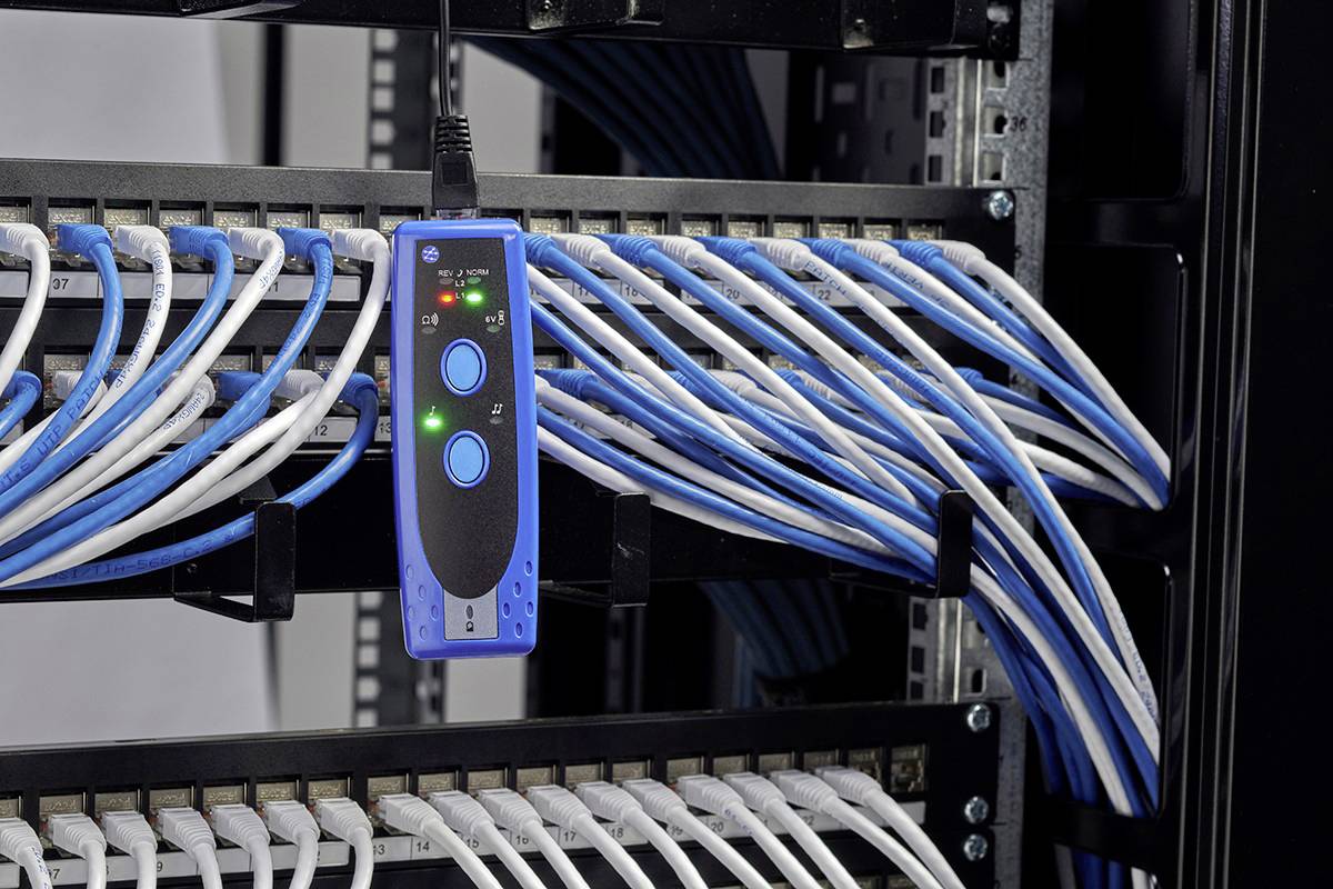 Serverový rack s připojenými modrými a bílými ethernetovými kabely a kabelovým testerem s indikačními diodami visícím na svazku kabelů.