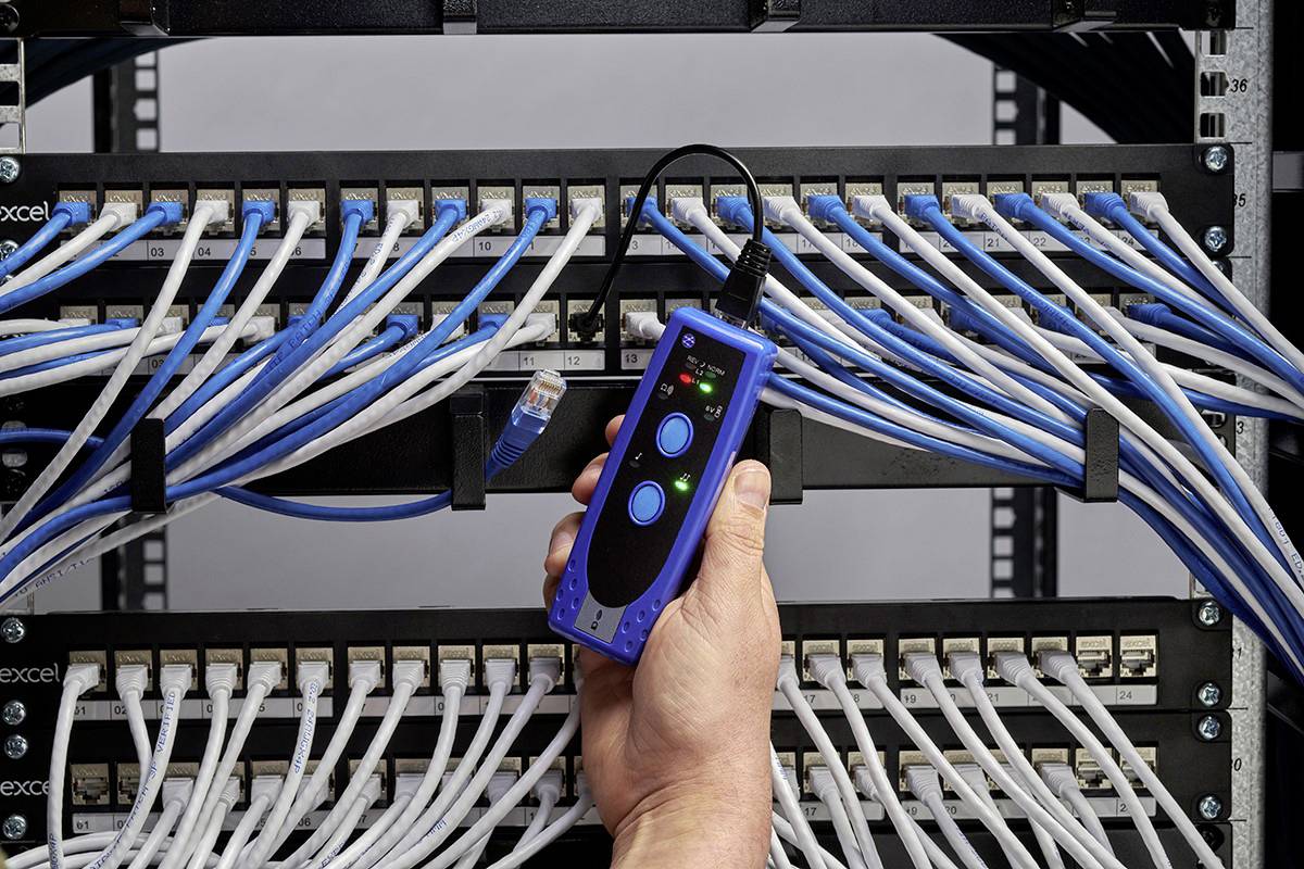 Osoba drží síťový tester nad patch panelem s mnoha modrými a bílými ethernetovými kabely.